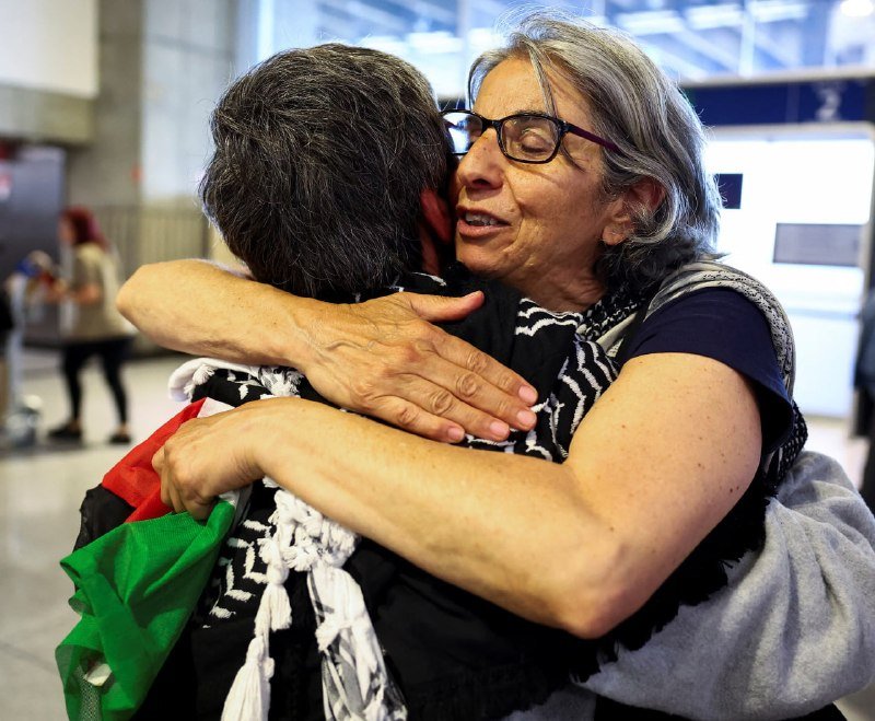 Two women embrace tightly in a public indoor space, with one holding a green and red bag while blurred figures and architectural structures appear in the background.