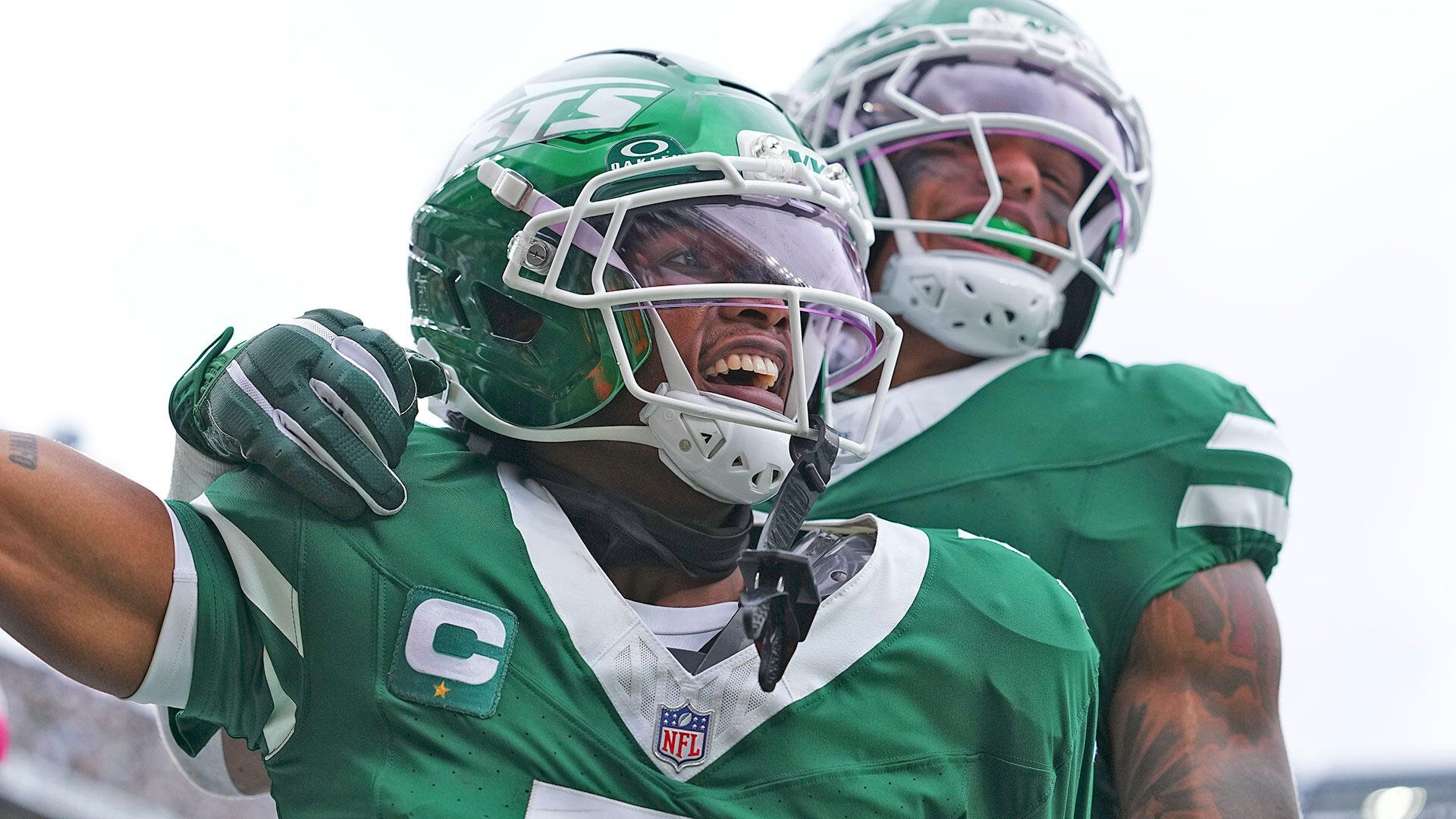 Two football players in green jerseys and helmets celebrate, with one wearing a captain's "C" patch, visibly cheering during an NFL game.