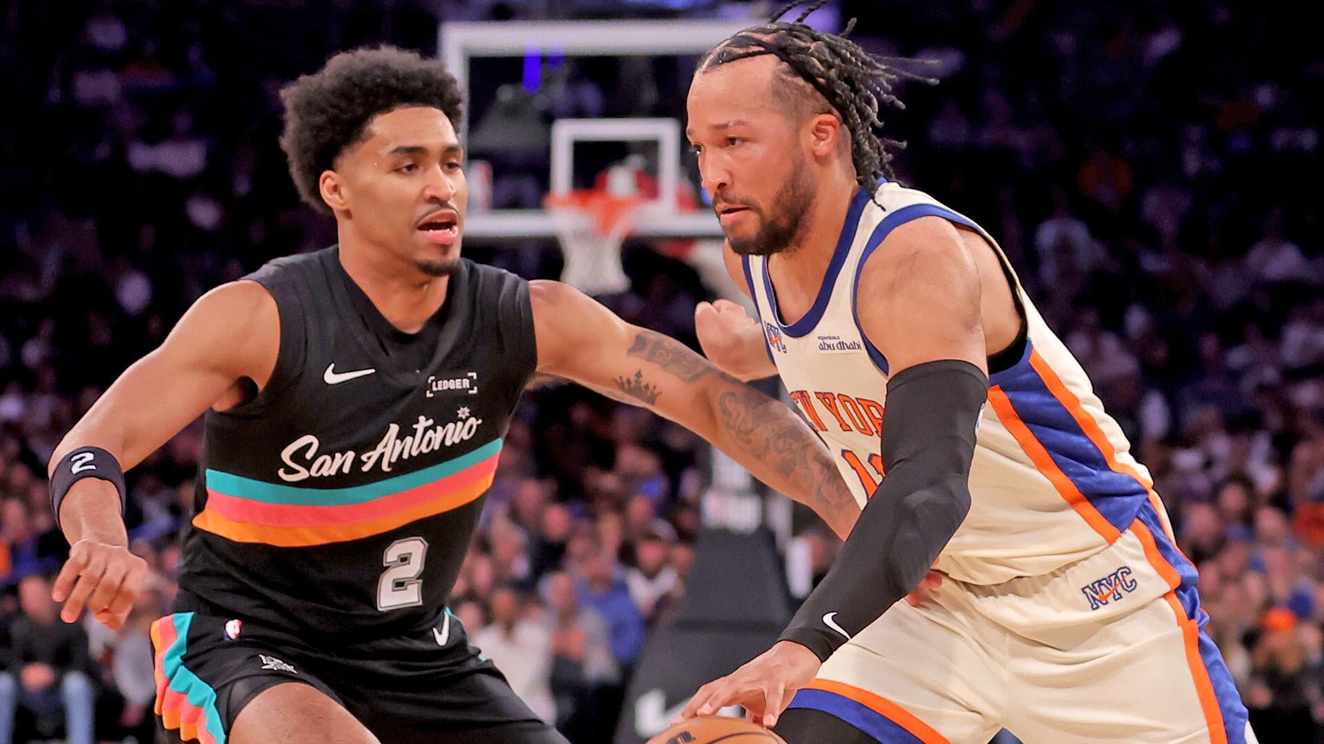 A San Antonio Spurs player in a black jersey defends against a New York Knicks player in a white jersey dribbling the ball during a basketball game.