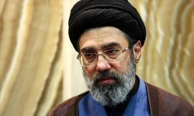 A bearded man wearing glasses, a black turban, and a brown robe over a blue garment looks toward the camera against a beige curtain backdrop.