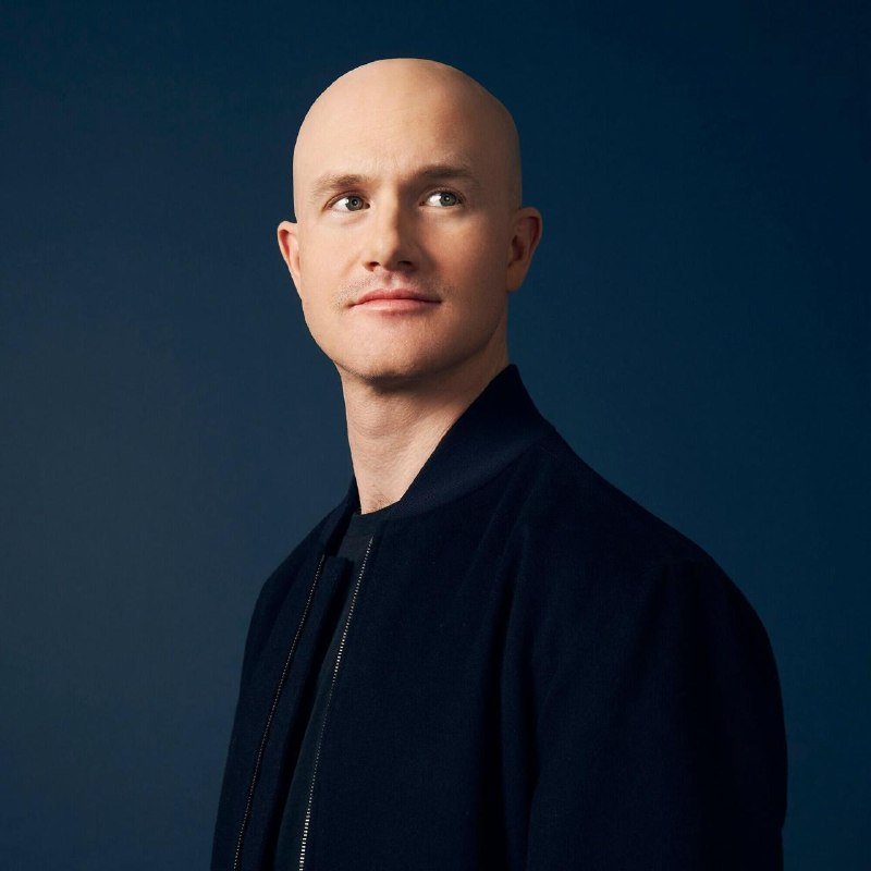 A bald man in a dark bomber jacket looks toward the camera against a navy blue background.