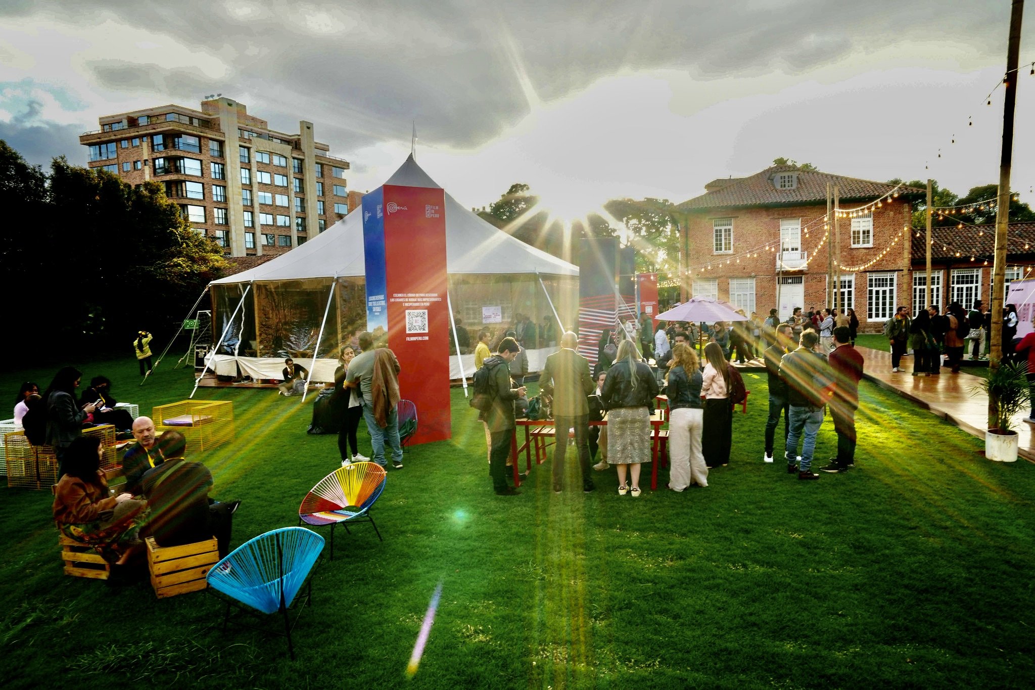 A crowd gathers on a grassy lawn at sunset near a white tent, a red promotional banner, and a brick building strung with lights.