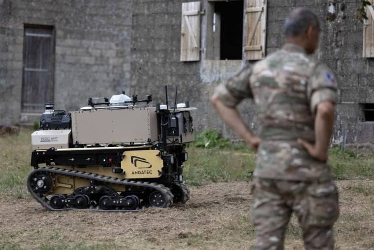 A tracked military robot labeled "Vigilant" and "AMGATEC" sits on grass in front of a weathered building while a person in camouflage uniform stands nearby.