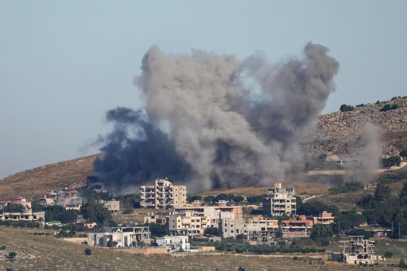 Large plumes of gray smoke rise from behind a cluster of hillside residential buildings against a clear blue sky.