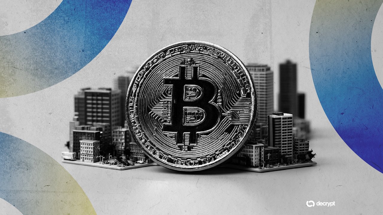 A large physical Bitcoin token stands upright in front of a miniature grayscale cityscape, with a "decrypt" logo in the corner.