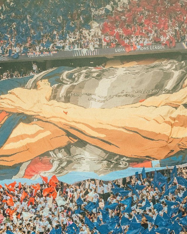 A stadium crowd waves blue and red flags while holding up a large banner displaying the UEFA Champions League trophy beneath signage reading "UEFA Champions League."