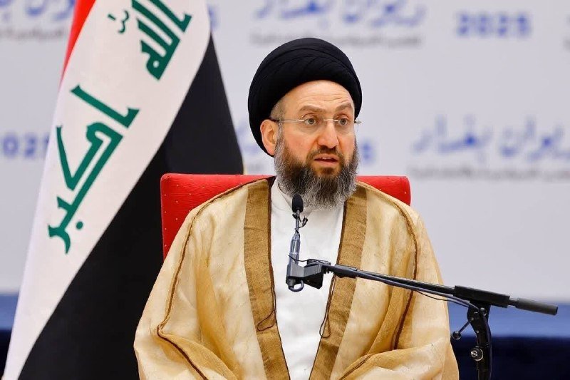 A bearded man wearing a black turban and gold-trimmed robe sits before a microphone beside an Iraqi flag.