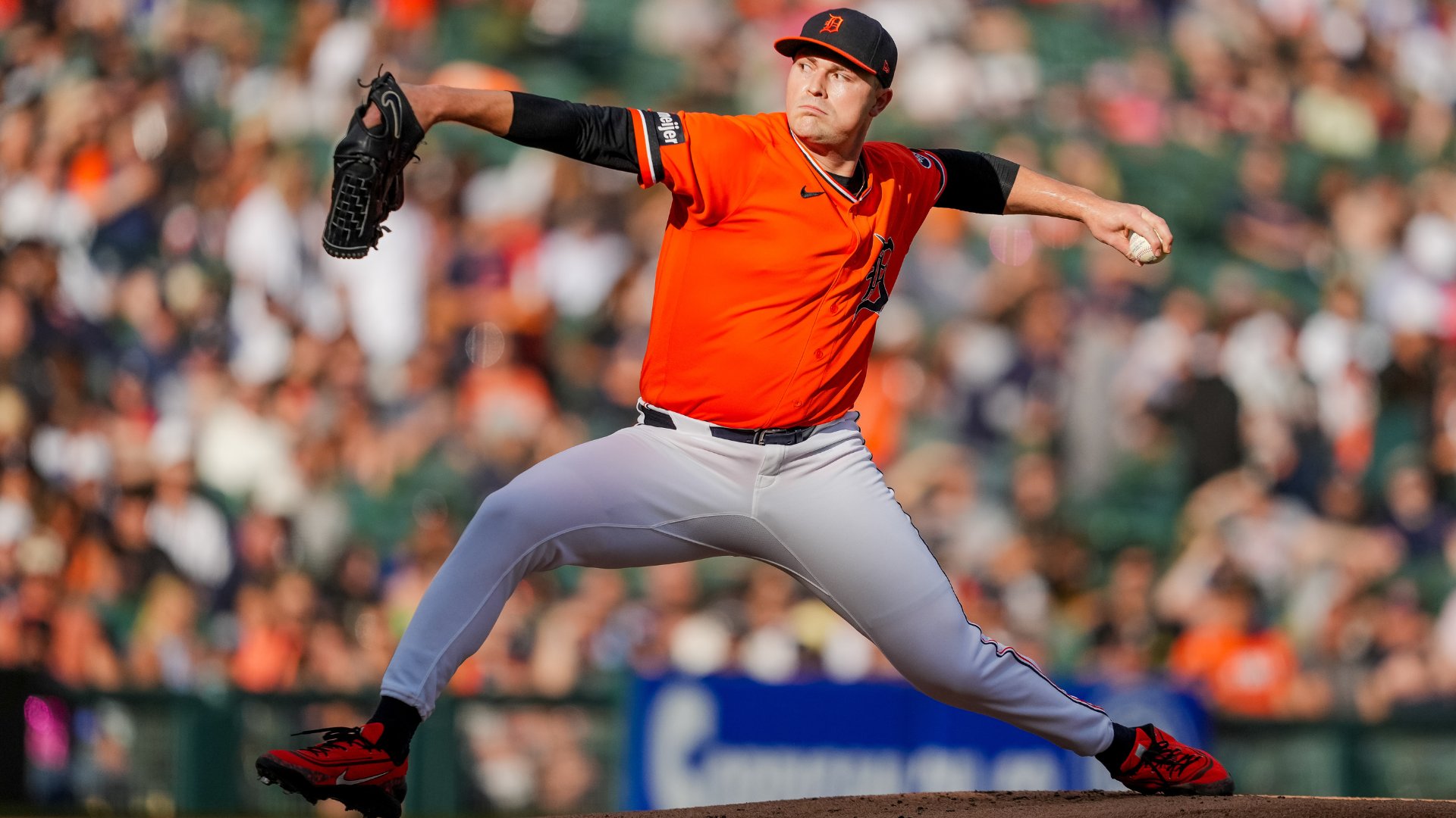 A baseball pitcher in an orange jersey and navy cap throws a pitch, with a blurred crowd in the background.