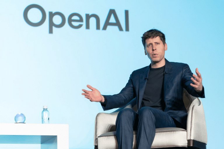 A man in a navy suit gestures with both hands while seated in a beige armchair on stage, with a large "OpenAI" logo displayed on the screen behind him.