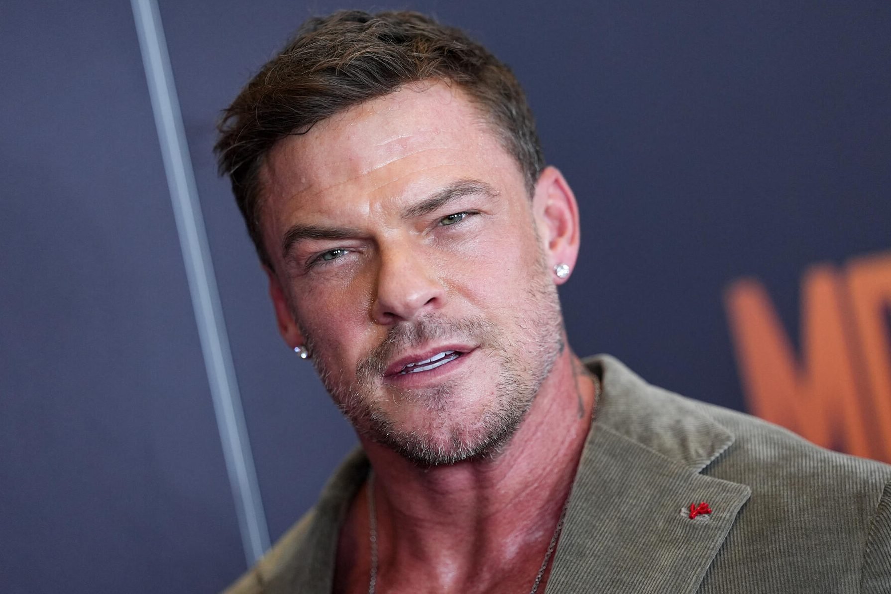 A close-up photograph shows a muscular man with short brown hair, stubble, and diamond earrings, wearing a tan corduroy jacket with a red lapel pin.
