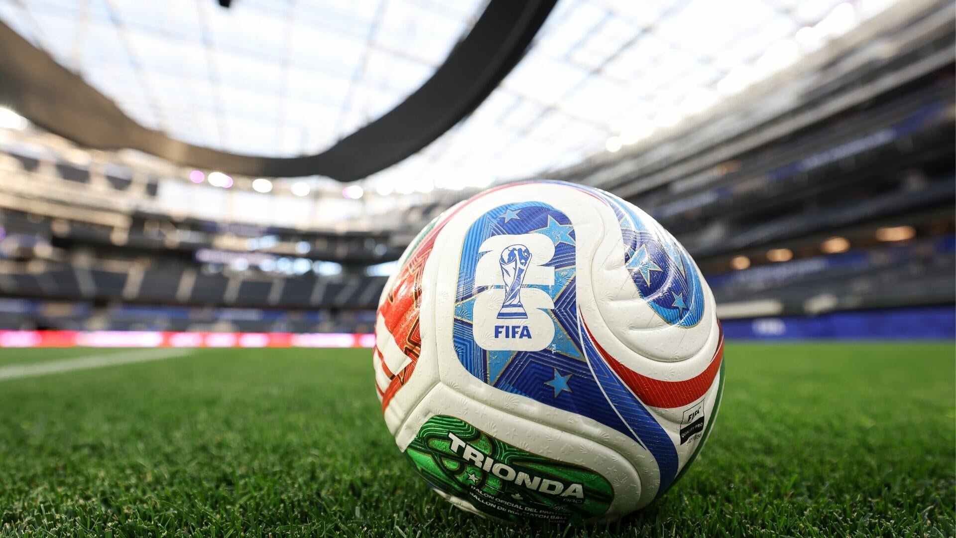 A white, blue, and red soccer ball branded "TRIONDA" with a "FIFA" logo sits on green grass inside a large, empty stadium with tiered seating.