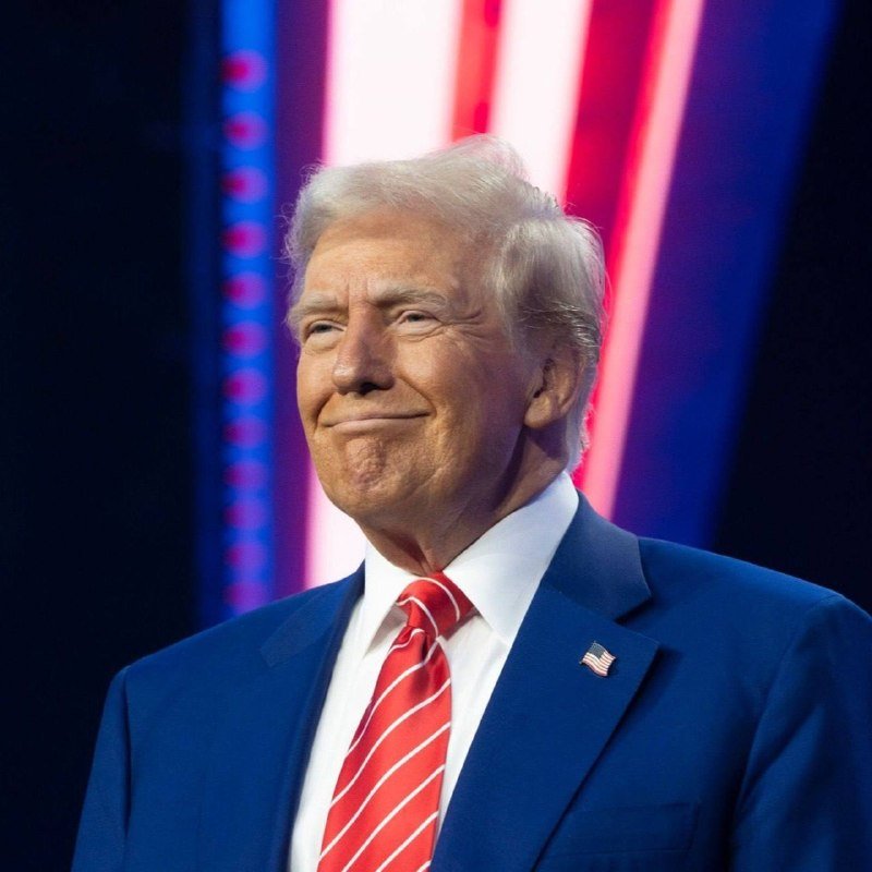 An older man with blonde hair wearing a dark blue suit, red striped tie, and an American flag pin smiles slightly against a backdrop of red, white, and blue illuminated vertical stripes.