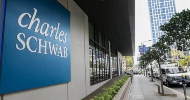 A blue Charles Schwab sign hangs on a building exterior along an urban sidewalk, with tall office buildings, trees, and parked vehicles visible down the street.