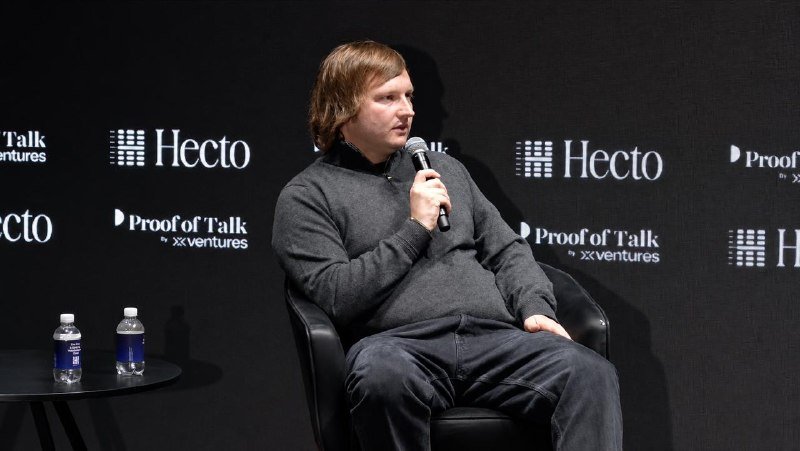 A man with shoulder-length hair, wearing a gray sweater, speaks into a microphone while seated on stage at the "Proof of Talk by XVentures" event sponsored by Hecto.