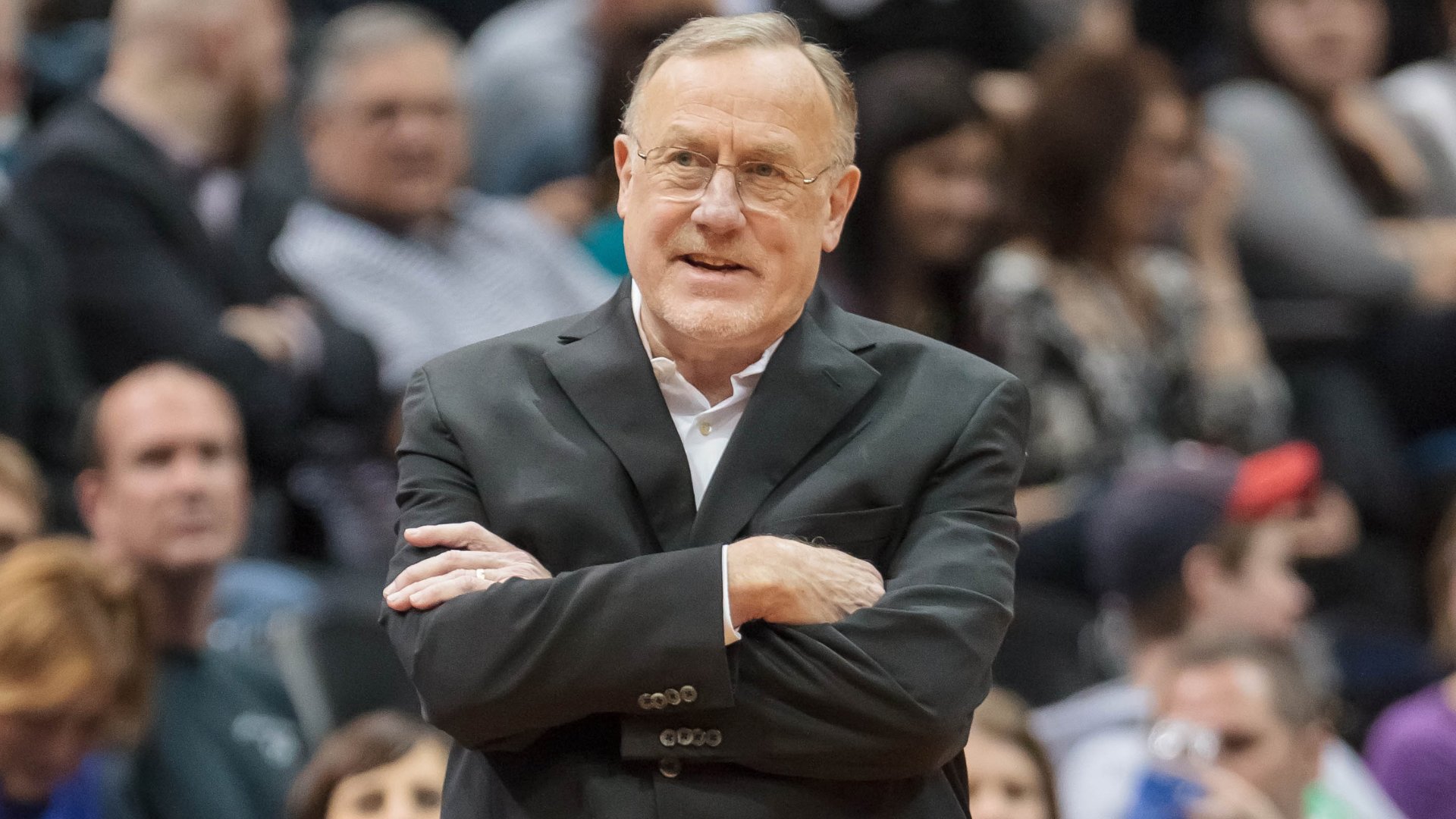 A man in a dark suit and glasses stands with crossed arms, smiling slightly, in front of a blurred crowd.