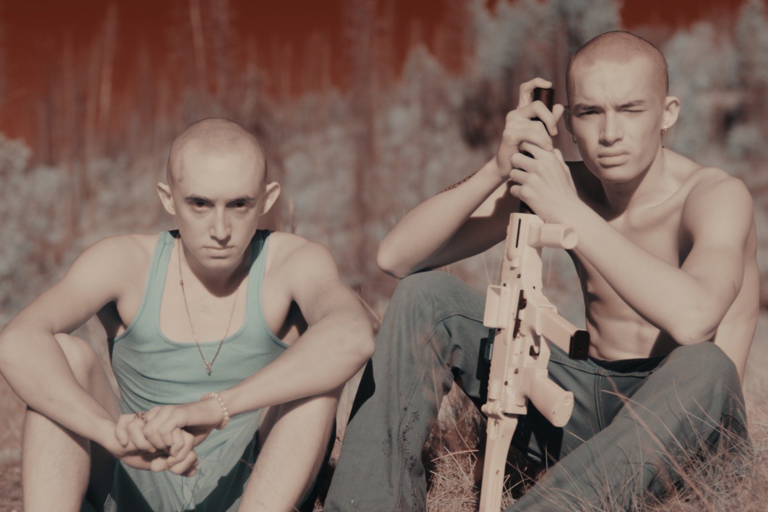 Two young men with shaved heads sit outdoors on dry grass against a rusted wall, one holding a tan rifle while wearing a tank top and chain.