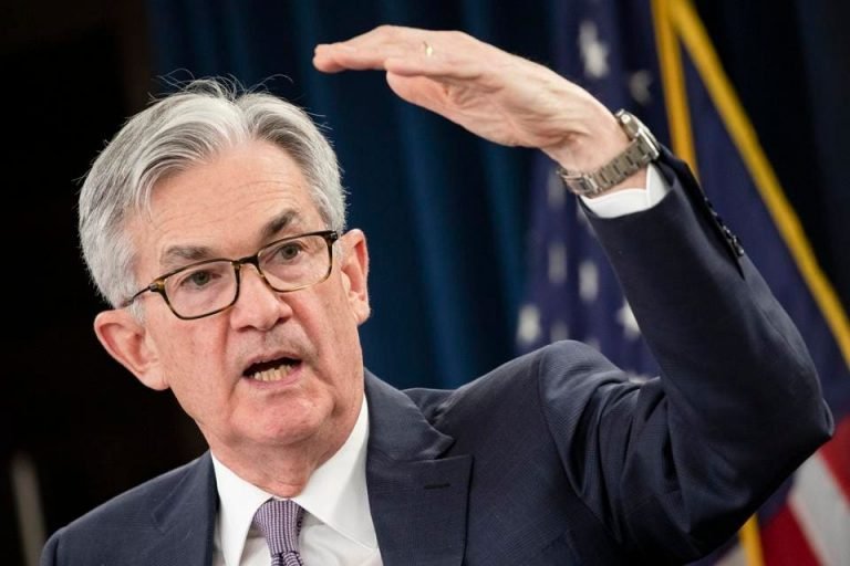 A gray-haired man in a suit and glasses gestures with his raised hand while speaking in front of an American flag.