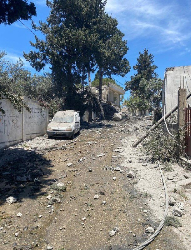 A debris-covered street with a dust-coated parked car, damaged buildings, fallen branches, and downed wires under a clear blue sky.