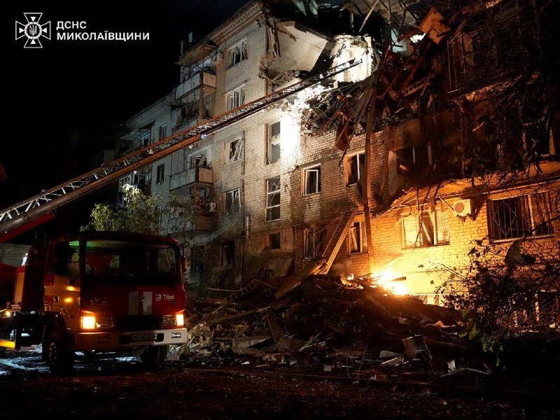 A fire truck with an extended ladder operates at night beside a heavily damaged multi-story brick apartment building with shattered windows and debris scattered on the ground.
