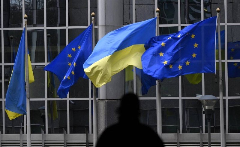 Multiple blue-and-yellow flags, including EU and Ukrainian designs, fly on flagpoles outside a glass-fronted building as a silhouetted figure stands in the foreground.