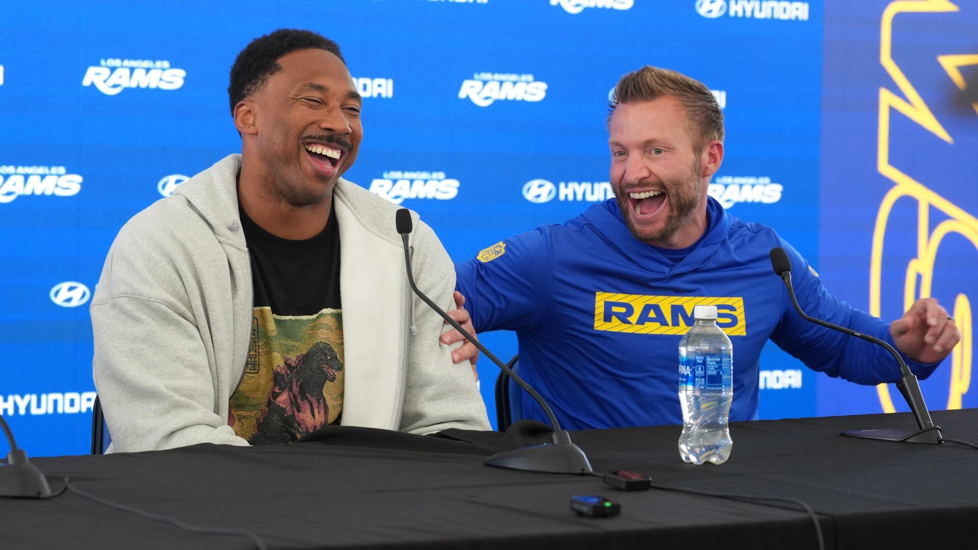 Two men laugh while seated at a press conference table with microphones, against a backdrop displaying Los Angeles Rams and Hyundai logos.