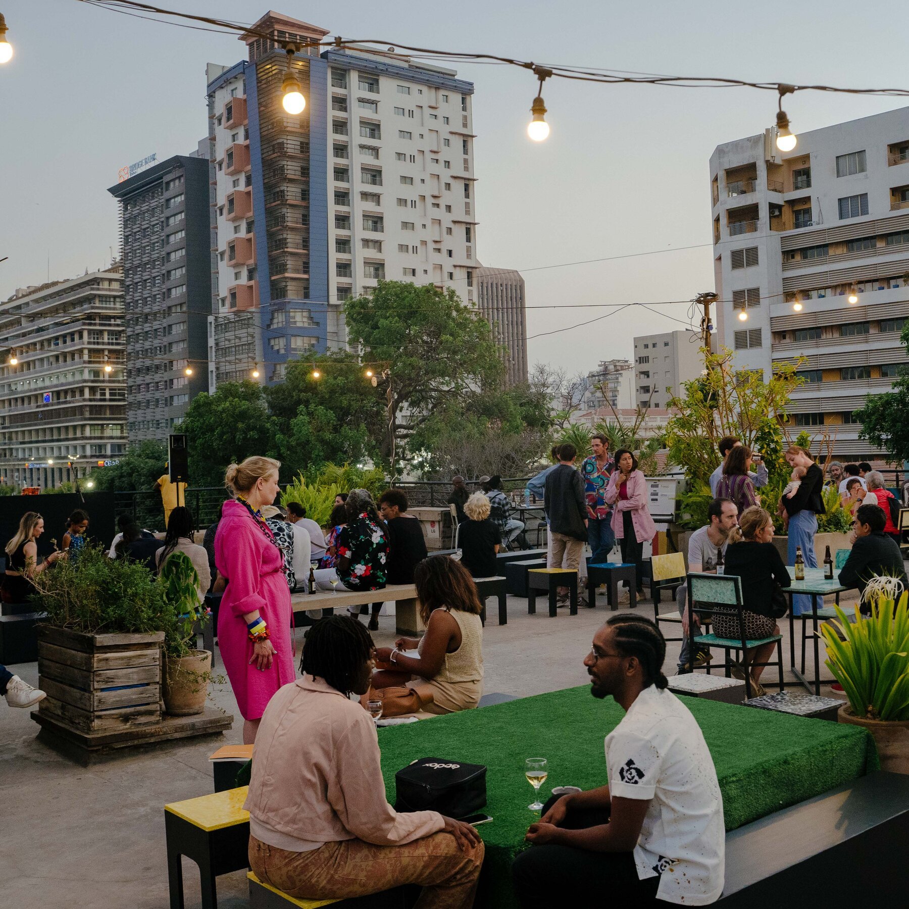 People gather at an outdoor rooftop venue with string lights, potted plants, and seating, set against a backdrop of tall city buildings at dusk.
