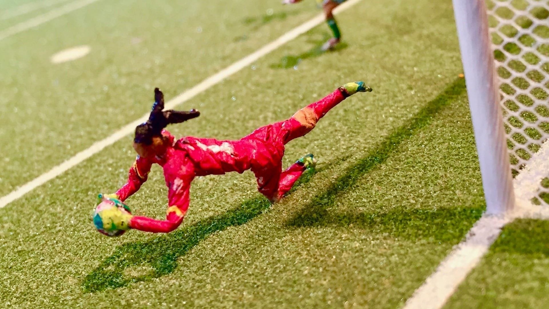 A goalkeeper in a red uniform dives to catch a yellow and green ball near the goalpost on a grass soccer field.
