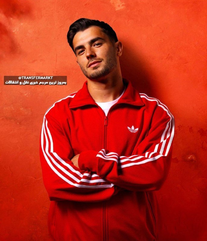 A dark-haired man in a red Adidas track jacket stands with crossed arms against a textured orange wall.