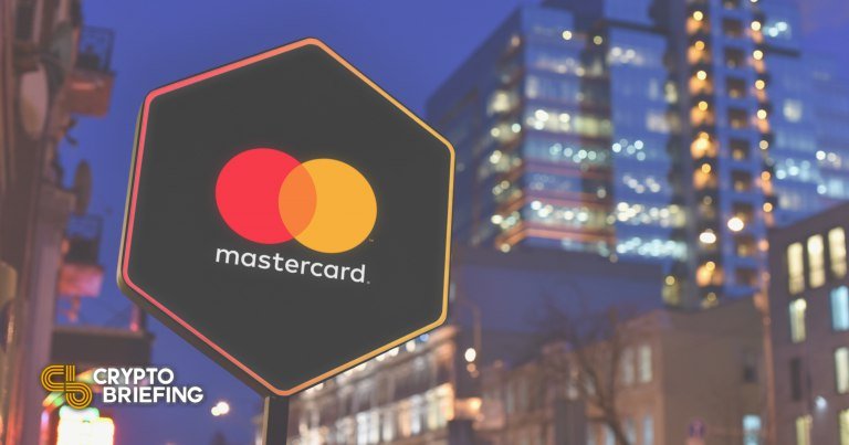 An illuminated hexagonal sign displaying the red and yellow Mastercard logo stands against a backdrop of lit high-rise buildings at dusk.