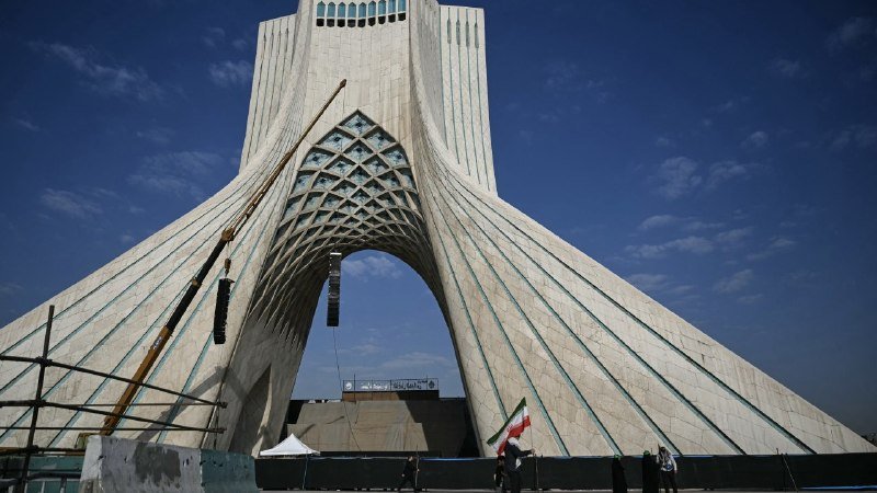 A towering white monument with a large arch and intricate tilework stands against a blue sky, with a person holding an Iranian flag at its base.
