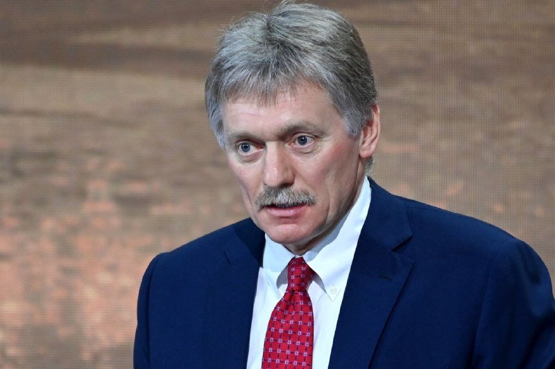 A gray-haired man with a mustache, wearing a navy suit and red patterned tie, looks downward against a blurred backdrop.