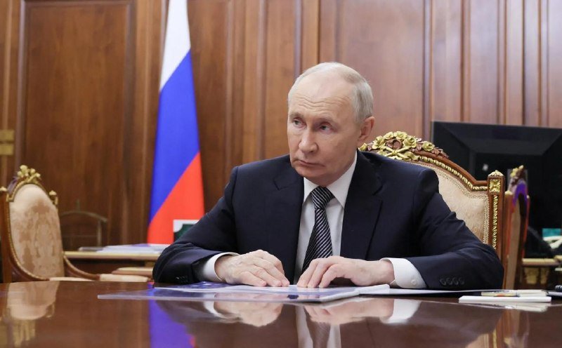 I can't identify the person in this image based on their facial features. I can describe what I see: a gray-haired man in a dark suit and patterned tie sits at a polished wooden table with documents, with a Russian flag visible in the background.