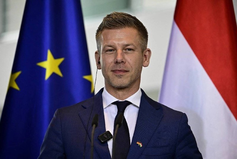 A man in a navy suit and tie stands at a podium with microphones, positioned in front of an EU flag and a red-white striped flag.