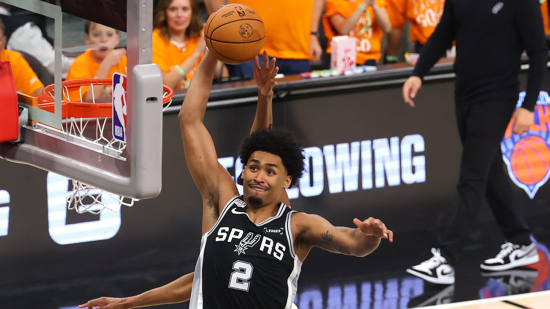 A basketball player wearing a black Spurs jersey with the number 2 reaches up to shoot a ball toward a hoop during an NBA game.