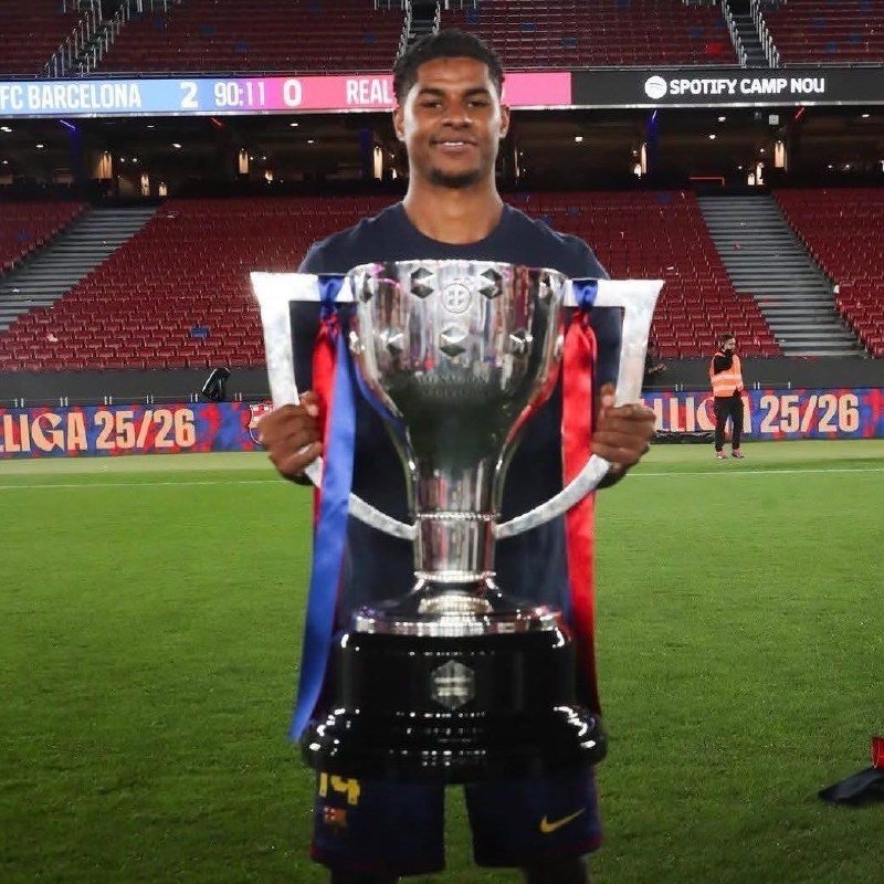 A smiling soccer player holds a large silver La Liga trophy on the field at an empty stadium displaying "FC BARCELONA 2 - 0 REAL" and "SPOTIFY CAMP NOU."