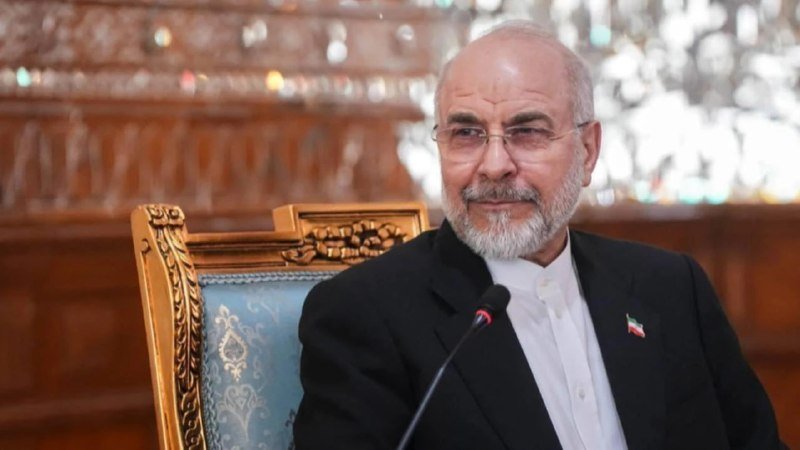 A bearded man in a dark suit and white shirt sits before a microphone in an ornately decorated room with chandeliers and wood paneling.