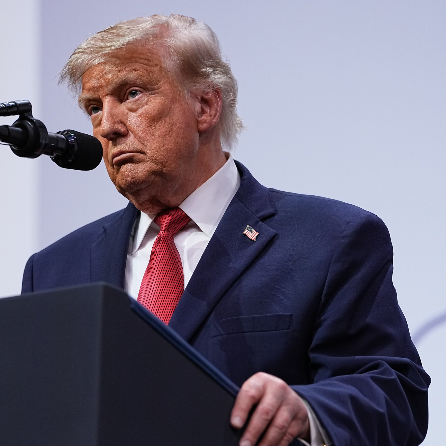 An older man with white-blond hair, wearing a navy suit, red tie, and an American flag lapel pin, stands at a podium beside a microphone.