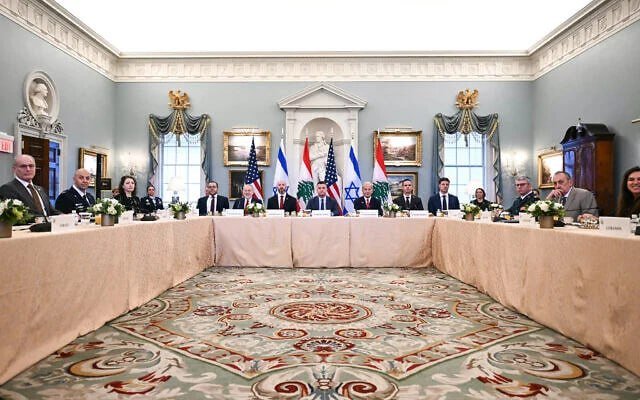 Officials sit around a U-shaped table with U.S. and another nation's flags displayed behind them in an ornate, blue-walled meeting room.