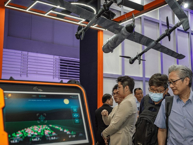 Several attendees view a suspended drone model and an illuminated display screen at an indoor exhibition booth.