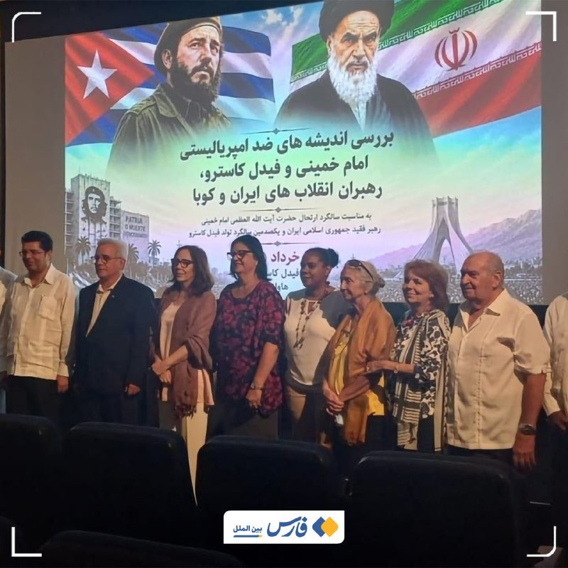A group of people stand together in an auditorium in front of a large screen displaying portraits of two men alongside Cuban and Iranian flags with Persian text.