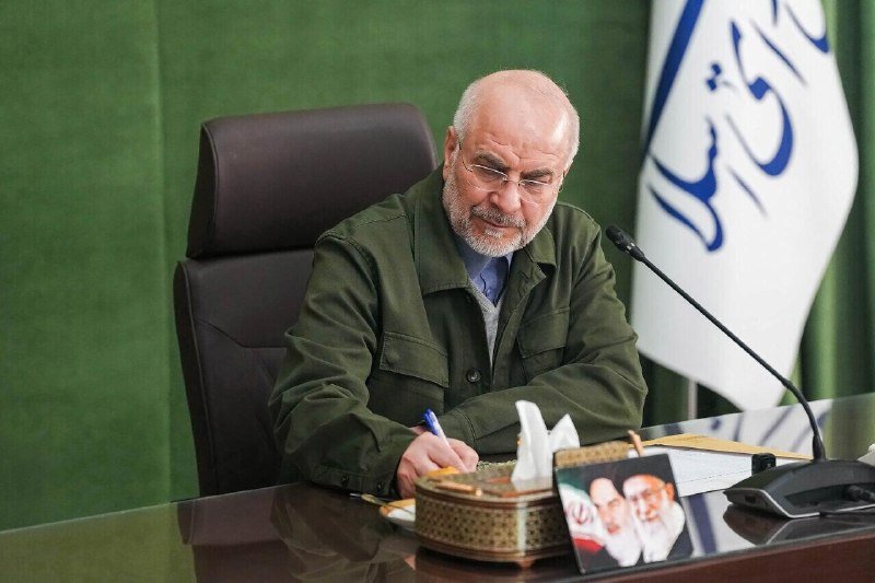 A bearded man in a green jacket sits at a desk writing with a pen, a microphone in front of him and an Iranian flag visible in the background.