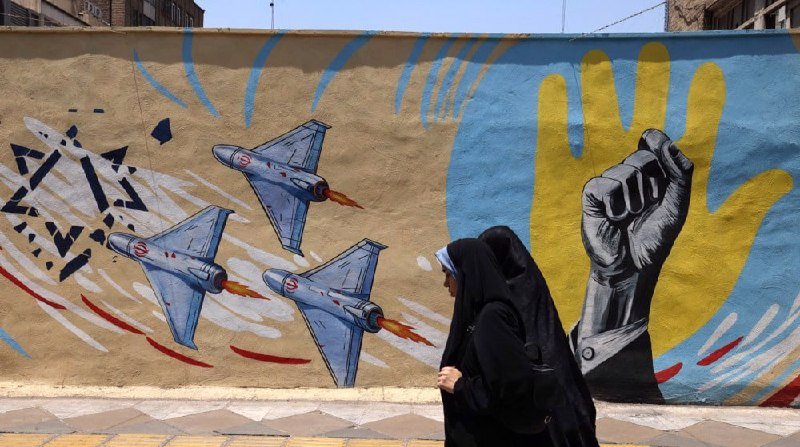 Two women in black chadors walk past a mural depicting fighter jets, a raised fist, and a yellow hand against a blue and beige wall.