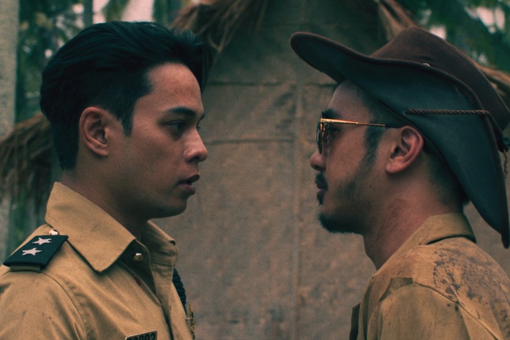 Two men in tan uniforms stand face-to-face outdoors; the left has star insignia on his collar, the right wears a wide-brimmed hat and sunglasses.