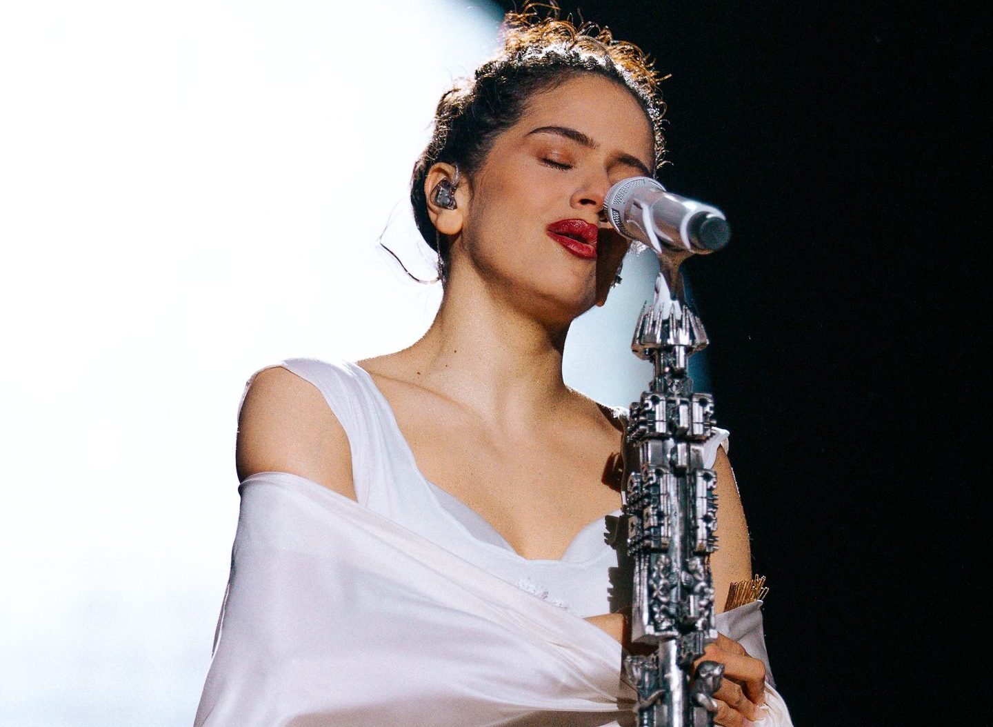 A woman with dark hair pulled back sings into a microphone on an ornate silver stand, eyes closed, wearing a draped white garment.