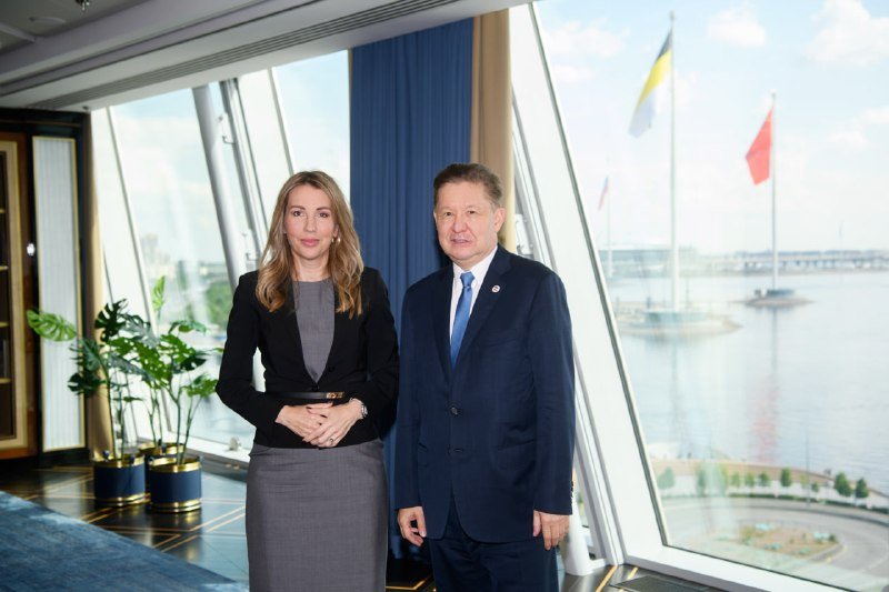 A woman in a gray dress and black blazer stands beside a man in a dark suit beside large windows overlooking a river with flags flying on distant boats.