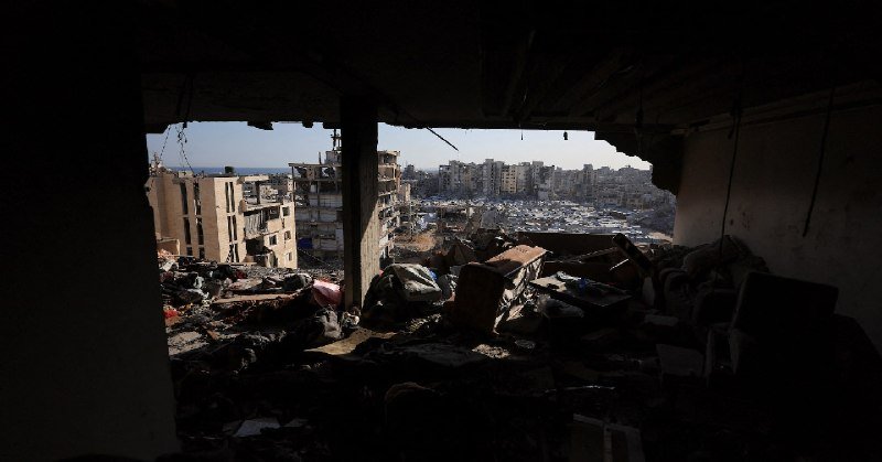Debris-filled interior of a damaged building overlooks a cityscape of densely packed, weathered apartment blocks under a clear sky.