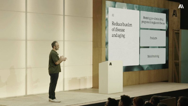 A man in a green shirt gestures while presenting on stage beside a large screen displaying "Reduce burden of disease and aging."