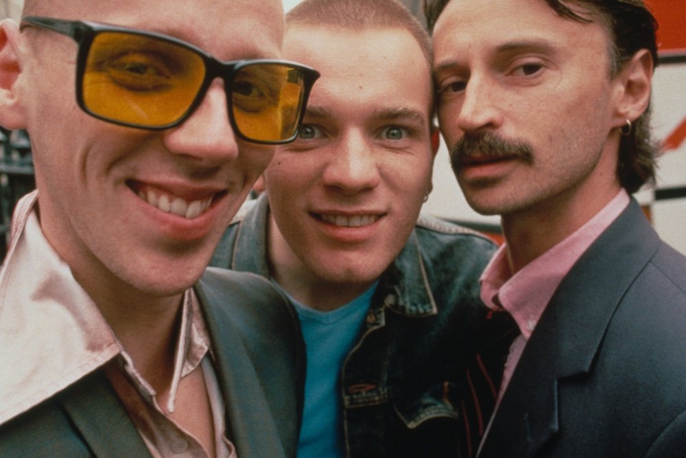 Three men pose closely together for a photograph: one wearing yellow-tinted sunglasses on the left, a smiling man in a denim jacket and blue shirt in the center, and a mustached man in a dark blazer on the right.