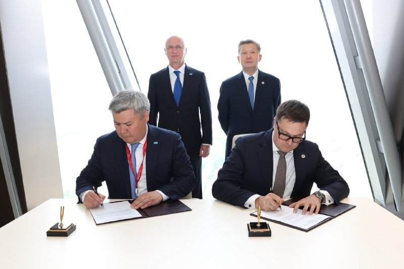 Two men in suits sign documents at a table while two other men in suits stand behind them in a room with angled windows.
