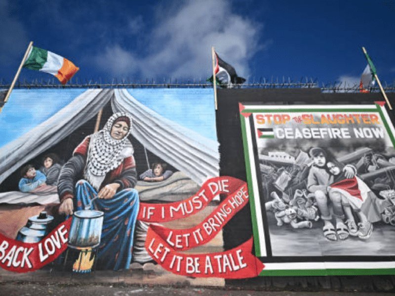 A mural features a woman with children beside a tent, alongside a "STOP THE SLAUGHTER CEASEFIRE NOW" poster, topped by Irish and Palestinian flags under a blue sky.